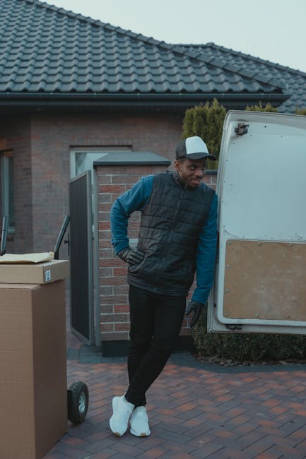 A man dressed in a blue long-sleeve shirt, black padded vest, and black trousers stands outside a residential property during a home relocation, leaning against the back of a white moving van with a wooden panel on the side. The van is parked on a brick-paved driveway adjacent to a house with a tiled roof and a brick exterior wall. Nearby, there are several packed cardboard boxes, some on a hand trolley, ready for loading or unloading. The man is wearing white sneakers and black gloves, and appears to be taking a break or preparing to assist with furniture transport and packing during a furniture move. The scene is set in natural daylight, with the house door open in the background, part of the ongoing packing and moving process supported by Man with Van South Kensington, specializing in removals and home relocation services.