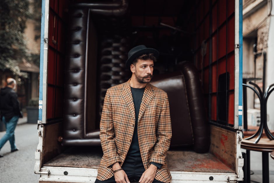 A man with dark hair, a beard, and wearing a black hat and a brown checkered blazer stands inside the back of a large moving van parked on a street, with its rear doors open. The van’s interior is lined with padded, leather-like dark brown materials, and the floor appears wooden. Behind the man, several large, wrapped furniture items are visible, including a sizeable dark brown armchair or sofa that is partially wrapped in protective plastic. To the left, outside the van, a person is walking away, dressed in casual clothing, with other buildings and pedestrians also visible in the background. The scene depicts the process of home relocation, showing furniture being prepared for transport as part of a professional removals service, possibly by Man with Van South Kensington, supporting efficient and careful furniture transport during a house move.