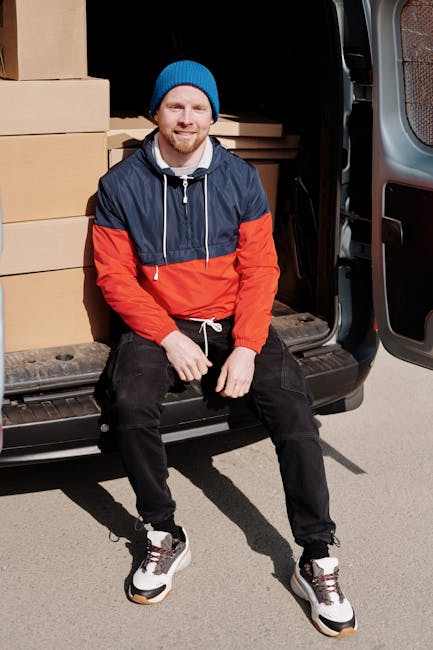 A man dressed in a blue long-sleeve shirt, black padded vest, and black trousers stands outside a residential property during a home relocation, leaning against the back of a white moving van with a wooden panel on the side. The van is parked on a brick-paved driveway adjacent to a house with a tiled roof and a brick exterior wall. Nearby, there are several packed cardboard boxes, some on a hand trolley, ready for loading or unloading. The man is wearing white sneakers and black gloves, and appears to be taking a break or preparing to assist with furniture transport and packing during a furniture move. The scene is set in natural daylight, with the house door open in the background, part of the ongoing packing and moving process supported by Man with Van South Kensington, specializing in removals and home relocation services.
