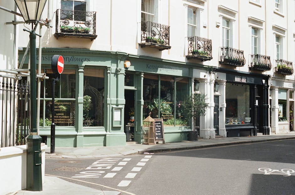 A street scene in Kensington and Chelsea, showing a row of retail shops with large glass windows and decorative wrought iron balconies above. The shop to the left has a pale green facade with signage indicating a flower shop and delivery services, and a small chalkboard sign outside. The shop to the right features a black storefront with white trim. A metal pole with a red and white no-entry sign is positioned near the pavement corner, and there is a pedestrian crossing with dashed white lines marked on the asphalt. The street scene is captured during daylight with natural lighting, and no people or vehicles are visible at this moment. This setting illustrates a suitable environment for home relocation or furniture transport services, with the available loading area and nearby parking regulations for removals, as provided by Man with Van South Kensington.