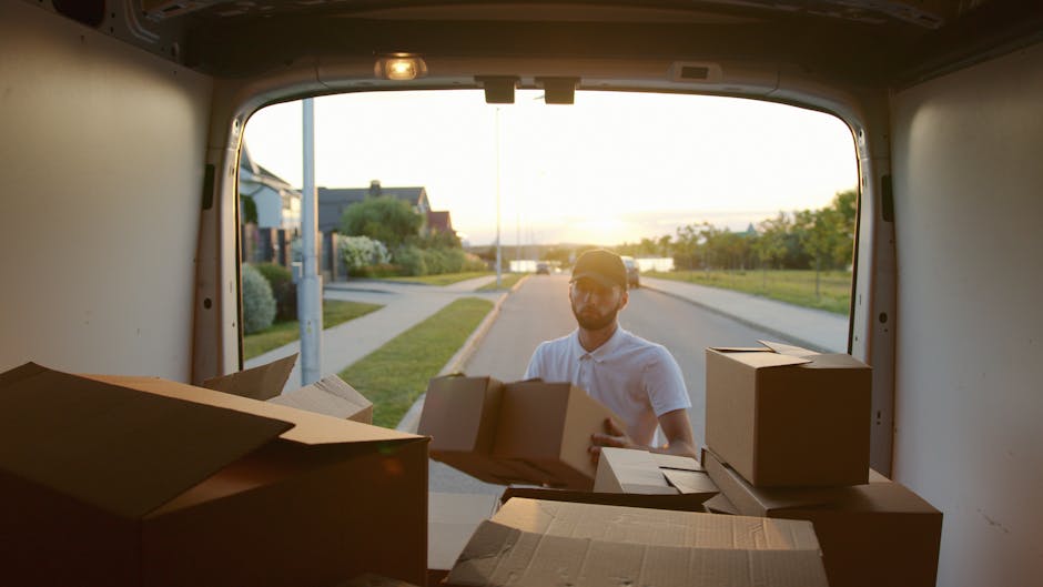 A man wearing a white shirt and a baseball cap is standing outside a white moving van during daylight, holding a cardboard box. The van's rear doors are open, revealing a partially packed interior filled with additional cardboard boxes, some stacked on top of each other. The scene shows a residential neighbourhood with houses, a streetlamp, greenery, and a sunset sky in the background, indicating home relocation or furniture transport process. The flooring inside the van is visible, and the boxes are arranged on the floor, prepared for loading or unloading, with some additional packaging materials nearby. Man with Van South Kensington’s professional removals team appears to be coordinating the packing or moving operation, focusing on efficient logistics for house removals or packing and moving services in the Kensington and Chelsea area.