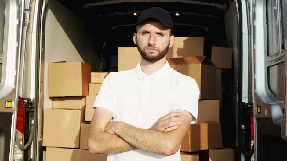 A young man with a beard, wearing a black cap and a white polo shirt, stands in front of an open moving van with his arms crossed. Inside the van, numerous cardboard boxes and packing materials are visible, arranged on the floor and against the sides of the vehicle. The boxes vary in size and are sealed with packing tape, designed for home relocation purposes. The van is parked outside a residential property, with the vehicle's interior illuminated by natural daylight. The loading area shows the process of furniture transport and packing during a house move, with the man representing a professional move coordinator or worker from Man with Van South Kensington. The backdrop suggests a typical urban or suburban setting suitable for home removals and logistics services.