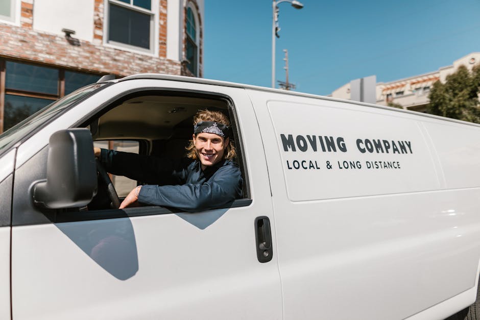 A smiling young man with long hair tied back and wearing a headband sits in the driver's seat of a white moving van, which has the words 'MOVING COMPANY LOCAL & LONG DISTANCE' printed on its side. The vehicle is parked outside a modern building with large windows, under a bright blue sky. The man is casually dressed in a dark top and appears to be preparing for a home relocation or furniture transport as part of a professional moving service, with the interior of the van visible through the open driver’s window. In the background, there are trees and other buildings, indicating an urban environment typical of house or office removals. The scene captures the loading process or departure phase of a furniture move, with the van positioned on a paved area, ready for moving logistics carried out by Man with Van South Kensington. This image emphasizes the professional moving process, including the vehicle, packing materials, and the staff involved in residential or commercial relocations.