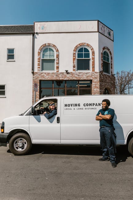 A white moving van with the words 'MOVING COMPANY LOCAL & LONG DISTANCE' printed on the side is parked in front of a two-storey building with a white and brick facade. Inside the van's cab, a man with long hair and sunglasses is seated, holding the steering wheel and looking out. Standing next to the van, another man with a beard, wearing a dark blue shirt and trousers, has his arms crossed and is smiling. The scene appears to be part of a home or office relocation process, with the van positioned on a paved street in a residential or commercial area. Behind them, the building features large ground-floor windows and arched upper-floor windows, with the sky clear and blue overhead. This setup suggests the beginning or continuation of furniture transport, packing, or loading activities typical of professional removals services at [DOMAIN].