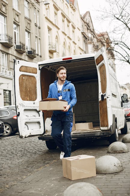 A man wearing a blue work uniform with a manwithvansouthkensington.com logo on the chest stands outside a white moving van with open rear doors, holding a cardboard box. The van is parked on a cobblestone street in South Kensington, near residential buildings with classic UK architecture, including balconies and sash windows. Several cardboard boxes, some wrapped in plastic or packing paper, are placed on the ground and inside the van, indicating a home relocation process. The scene captures the loading or unloading phase of furniture transport and packing during a house move, with the individual preparing to load or unload boxes, supported by equipment like trolleys and blankets for safe handling, against a backdrop of leafless trees and parked cars.