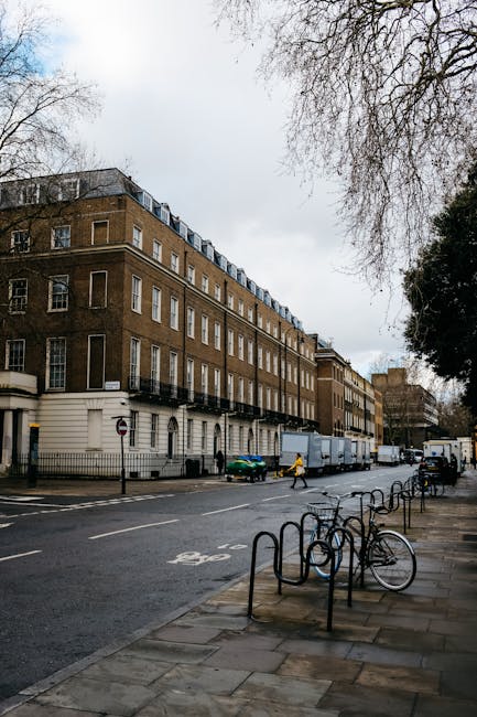 A wide exterior view of a multi-storey historic brick building on Old Brompton Road in South Kensington, SW7, with several windows and small balconies on each floor. Parked on the street in front of the building are delivery trucks and a small green car, with people walking nearby, including a person in a yellow jacket. The sidewalk features metal bike racks with a bicycle secured to one of them and is lined with leafless trees, suggesting late autumn or winter. The overcast sky above results in diffuse natural lighting, and part of a tree with bare branches is visible in the upper right corner. The scene captures the urban environment where house removals or furniture transport may take place, with the pavement and street prepared for loading and unloading activities, as facilitated by local moving service providers like Man with Van South Kensington.