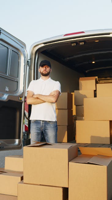 A man with a beard, wearing a dark cap, white t-shirt, and jeans, stands with arms crossed next to the open rear doors of a white van in an outdoor setting. The van's interior is visible, showing an organized load of large, cardboard moving boxes stacked neatly, some on the ground and others inside the vehicle. The boxes are plain brown with no visible markings. The scene depicts the loading process during a home relocation or furniture transport arranged by Man with Van South Kensington, with natural daylight illuminating the area, indicating an active packing and moving operation consistent with same-day removals services at South Kensington Gloucester Road.