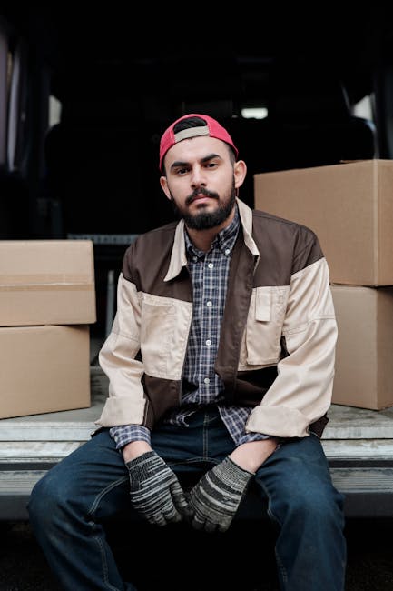 A young man with a beard and wearing a red cap, brown and beige jacket, checkered shirt, and striped gloves, sitting on the edge of a vehicle's open cargo area filled with stacked cardboard boxes, during a house removal or furniture transport process. The background shows the interior of the vehicle with a dark headrest and part of the vehicle's trunk visible. The setting suggests preparation for a home relocation or moving service, with the man appearing to rest after loading boxes inside the van, which is part of a professional removals operation by Man with Van South Kensington. The image emphasizes elements of packing and loading for a move, with the boxes positioned behind him on either side, indicating an organized moving process.