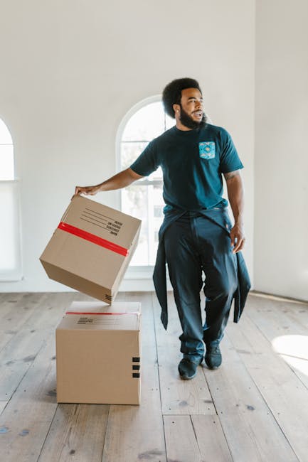 A man with dark curly hair, a beard, and dark skin, is inside a bright room with white walls and wooden flooring, holding a cardboard box with red tape in his right hand while standing next to two other boxed items. One box is on the floor, and another is being lifted. The room features large, arched windows allowing natural light to illuminate the space. The man is dressed in casual clothing, including a dark T-shirt, with a sweater tied around his waist. This scene depicts a home relocation or packing process, with the man preparing to load or unload boxes as part of furniture transport or moving services. The environment suggests a professional moving service such as Man with Van South Kensington, which is engaged in packing and moving activities as part of house removals for clients.
