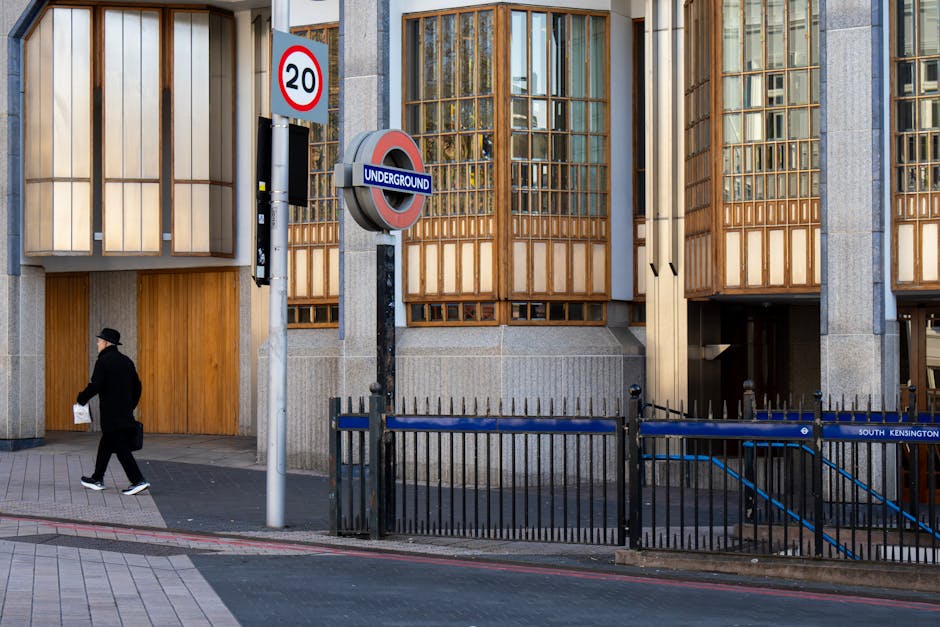 A man dressed in dark clothing, including a jacket, trousers, and a hat, is walking on the sidewalk near a modern building with wooden window panels and concrete walls. In the foreground, a black metal fence borders the pavement, with a blue and black striped cover over some kind of underground utilities or entrance. A sign indicating a 20 mph speed limit is mounted on a pole next to a circular underground station sign with red, blue, and white coloring, suggesting proximity to an underground metro station in South Kensington. The building's exterior features large windows with wooden framing, and the sidewalk is paved with light-colored tiles. Nearby, a parking area or loading zone is visible, consistent with an urban area near a house removal or relocation service. The scene appears to be during daytime with clear lighting, suitable for illustrating logistics and transport related to house removals or furniture transport, as handled by Man with Van South Kensington.